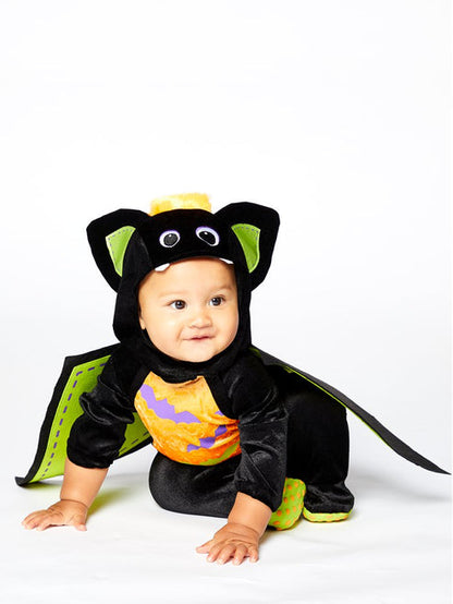 Baby wearing a plush black velvet bat romper with a bat-face hood and attached colorful felt wings in a playroom setting.
