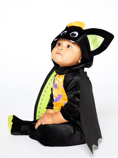 Baby wearing a plush black velvet bat romper with a bat-face hood and attached colorful felt wings in a playroom setting.