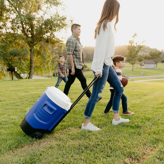 Side view of the rugged blue and white Igloo 5 Gallon Sport Rolling Cooler showing its large black wheels and recessed pouring spigot, resting on gravel at a sunny outdoor worksite.