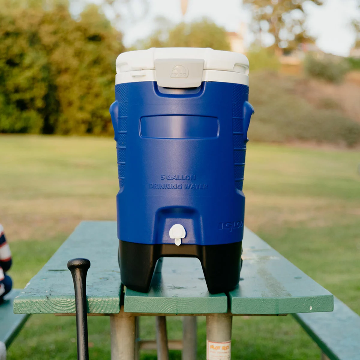 Side view of the rugged blue and white Igloo 5 Gallon Sport Rolling Cooler showing its large black wheels and recessed pouring spigot, resting on gravel at a sunny outdoor worksite.