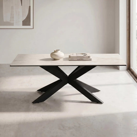 Sand-toned ceramic top rectangular coffee table with a bold black iron base, shown at a slight angle in front of a sofa in a bright living room, styled with books and a small vase.