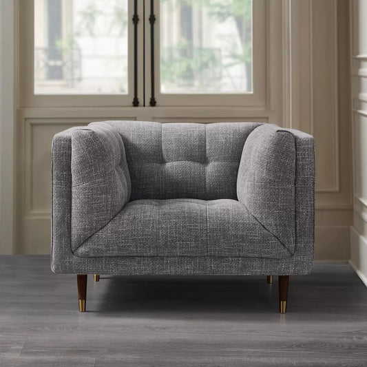 Front view of the Prospera Home Isla Grey Fabric Armchair showing hand-tufted ash grey upholstery and brass-tipped wooden legs in a brightly lit living room setting.