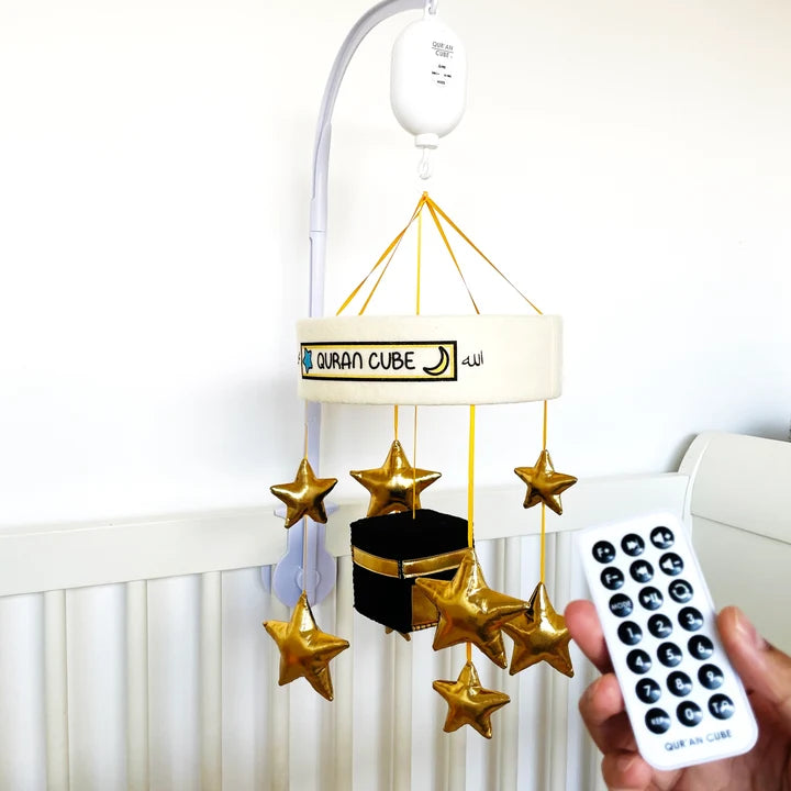 White baby cot mobile attached to a wooden crib rail featuring a plush central Kaaba surrounded by hanging golden stars and crescent moon plushies.
