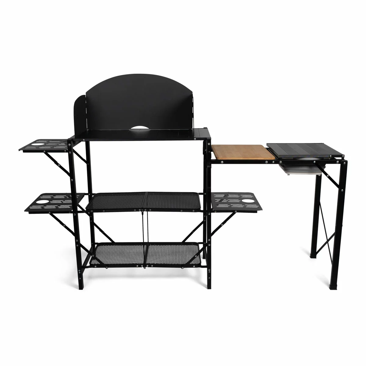 A charcoal grey Kampa Slim Chef Stand fully pitched on a grassy clearing, showing its metal windshield surrounding a camp stove, an integrated cutting board, and a popped-out green collapsible sink.