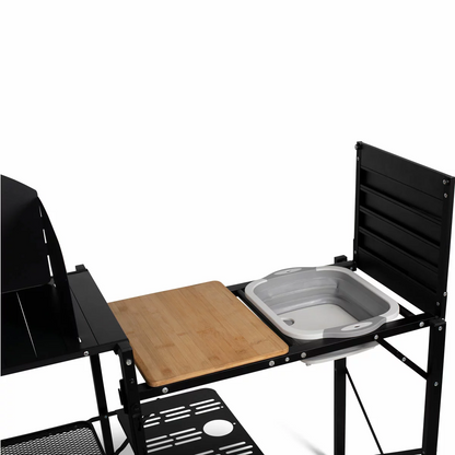 A charcoal grey Kampa Slim Chef Stand fully pitched on a grassy clearing, showing its metal windshield surrounding a camp stove, an integrated cutting board, and a popped-out green collapsible sink.