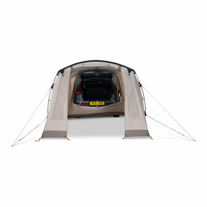A neutral beige Kampa Tailnest drive-away tent pitched on a grass campsite, with its rear tunnel attachment securely fitted over the open boot of a dark grey SUV vehicle.