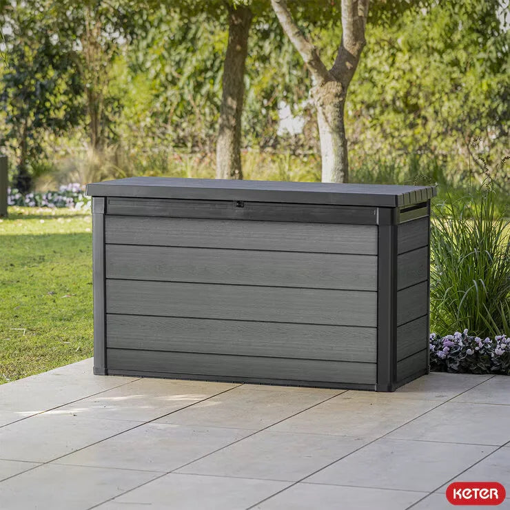 Grey Keter Cortina 757 litre outdoor storage deck box with realistic wood-effect Evotech finish and soft-opening lid positioned on a modern garden patio, storing outdoor cushions, garden tools, and sports equipment.