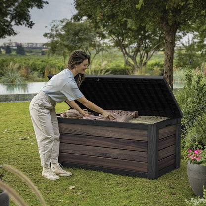 Front angle view of the brown Keter Rosewood 575L deck box featuring a realistic wood-grain texture, positioned on a modern stone patio next to outdoor green foliage.