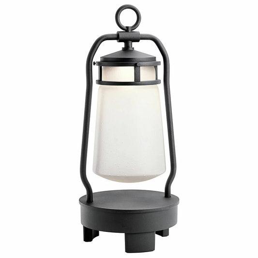 A textured black Kichler Lyndon Bluetooth speaker lantern with clear seeded glass emitting a warm glow on a dark patio table.