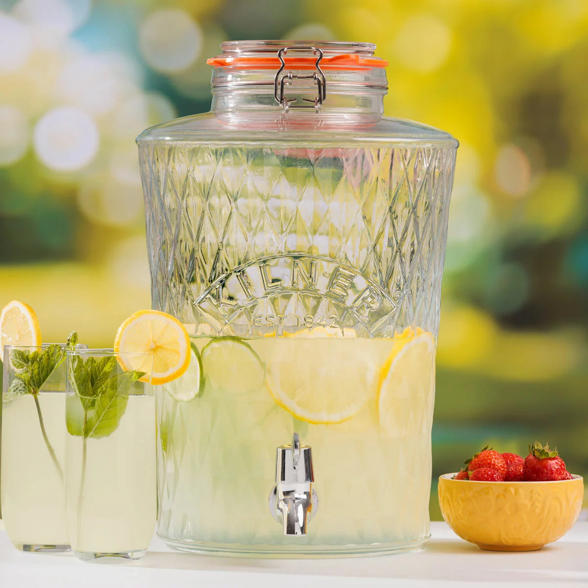 An 8-litre Kilner Diamond Drinks Dispenser filled with refreshing fruit-infused water, sitting on a wooden table, showcasing its faceted glass design and stainless steel clip-top.