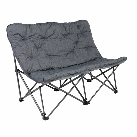 The KingCamp Oversized Padded Double Camping Chair in a neutral grey, set up on a grassy patch beside a lake, showcasing its padded backrest and robust steel frame.