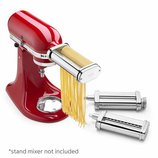 A silver KitchenAid 5KSMPRA pasta attachment set, featuring the roller and two cutters, neatly arranged on a clean kitchen counter next to a stand mixer.