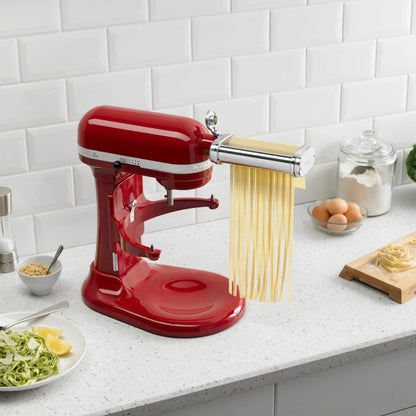 A silver KitchenAid 5KSMPRA pasta attachment set, featuring the roller and two cutters, neatly arranged on a clean kitchen counter next to a stand mixer.