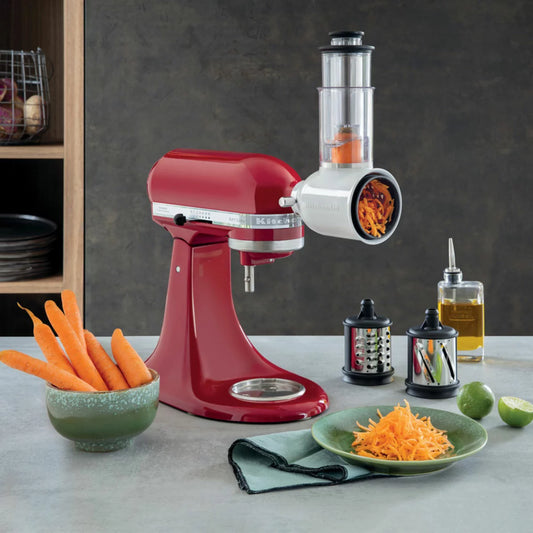 KitchenAid 5KSMVSA vegetable slicer and shredder attachment in silver, shown with its three stainless steel blade cylinders and storage case, sitting beside a stand mixer.