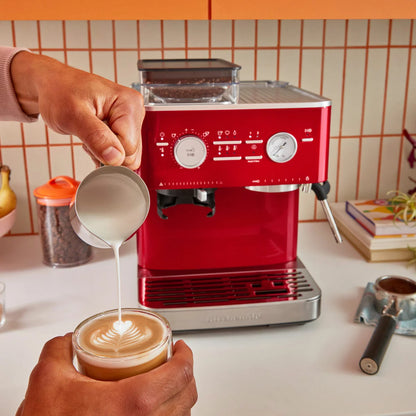 KitchenAid Candy Apple red semi automatic espresso machine with built in burr grinder, 58mm portafilter, and stainless steel milk pitcher on a clean kitchen countertop.