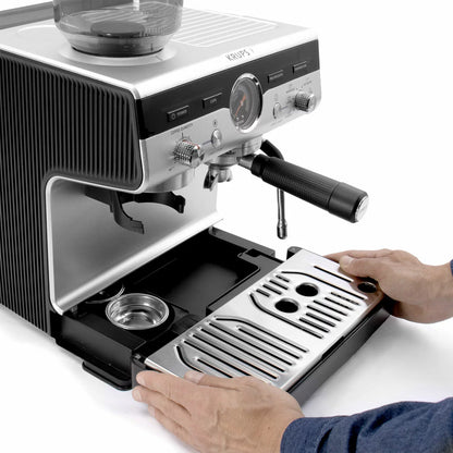 Front angle view of the black and stainless steel Krups XP801TG0 espresso machine showing the top bean hopper, portafilter attached to the group head, steam wand, and metal drip tray on a clean kitchen counter.