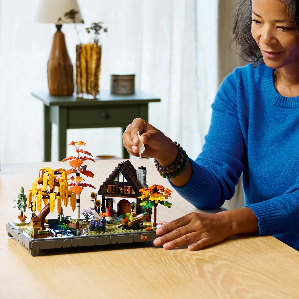 Three-quarter angle view of the LEGO Icons Autumn Cottage Garden set displaying orange maple trees, a stone stream, bird figures, and an openable cottage on a wooden table.