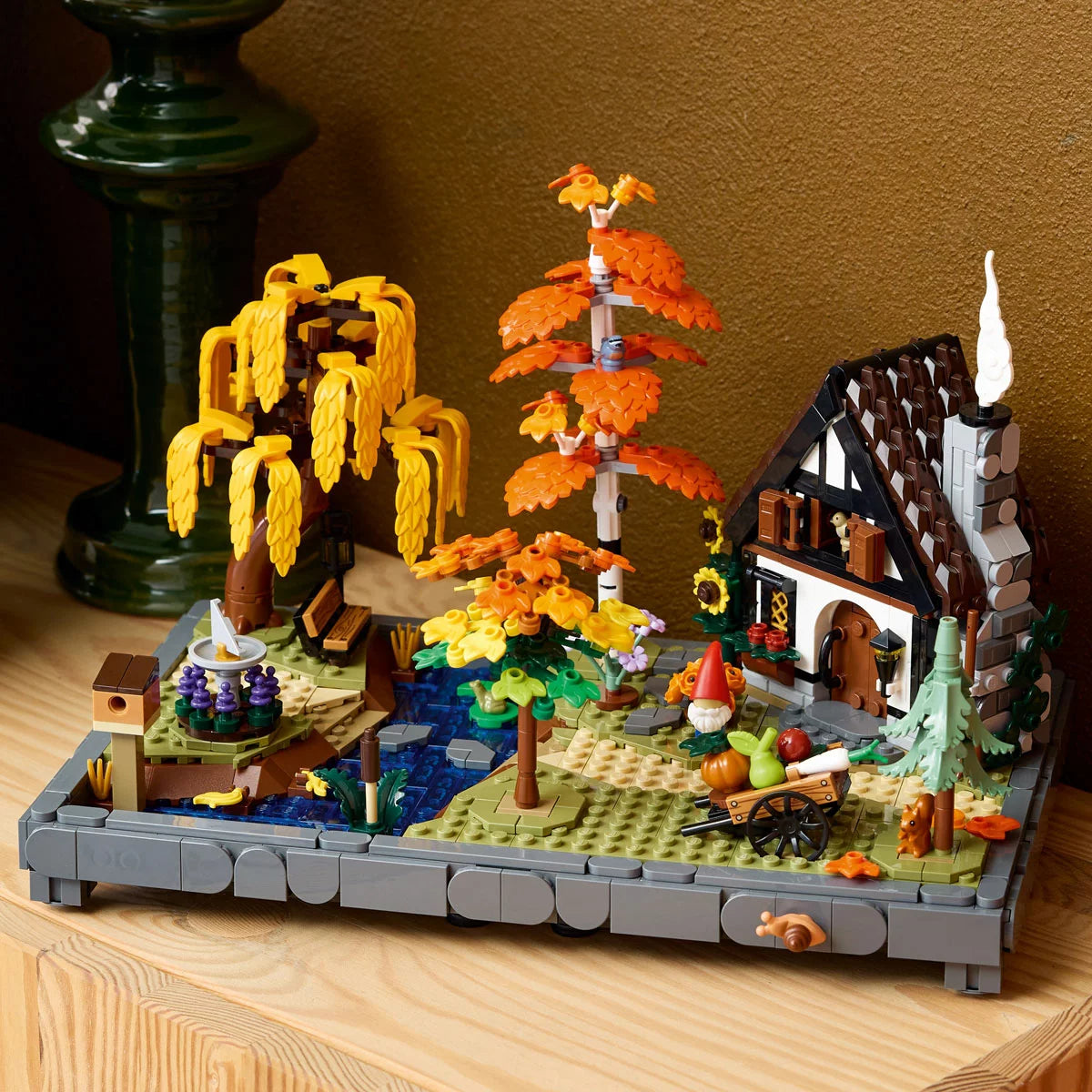 Three-quarter angle view of the LEGO Icons Autumn Cottage Garden set displaying orange maple trees, a stone stream, bird figures, and an openable cottage on a wooden table.