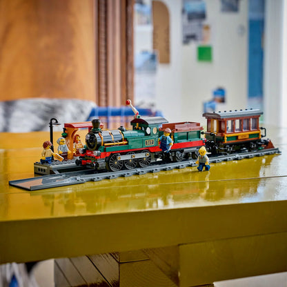Side profile view of the black LEGO City Vintage Steam Train with red wheels pulling a coal tender and passenger car along a section of track next to a station platform.