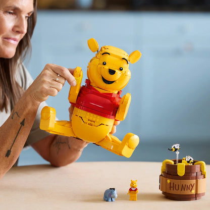 Three-quarter studio view of the built yellow LEGO 43300 Winnie the Pooh figure standing next to an open clay-colored honey pot revealing tiny interior rooms.
