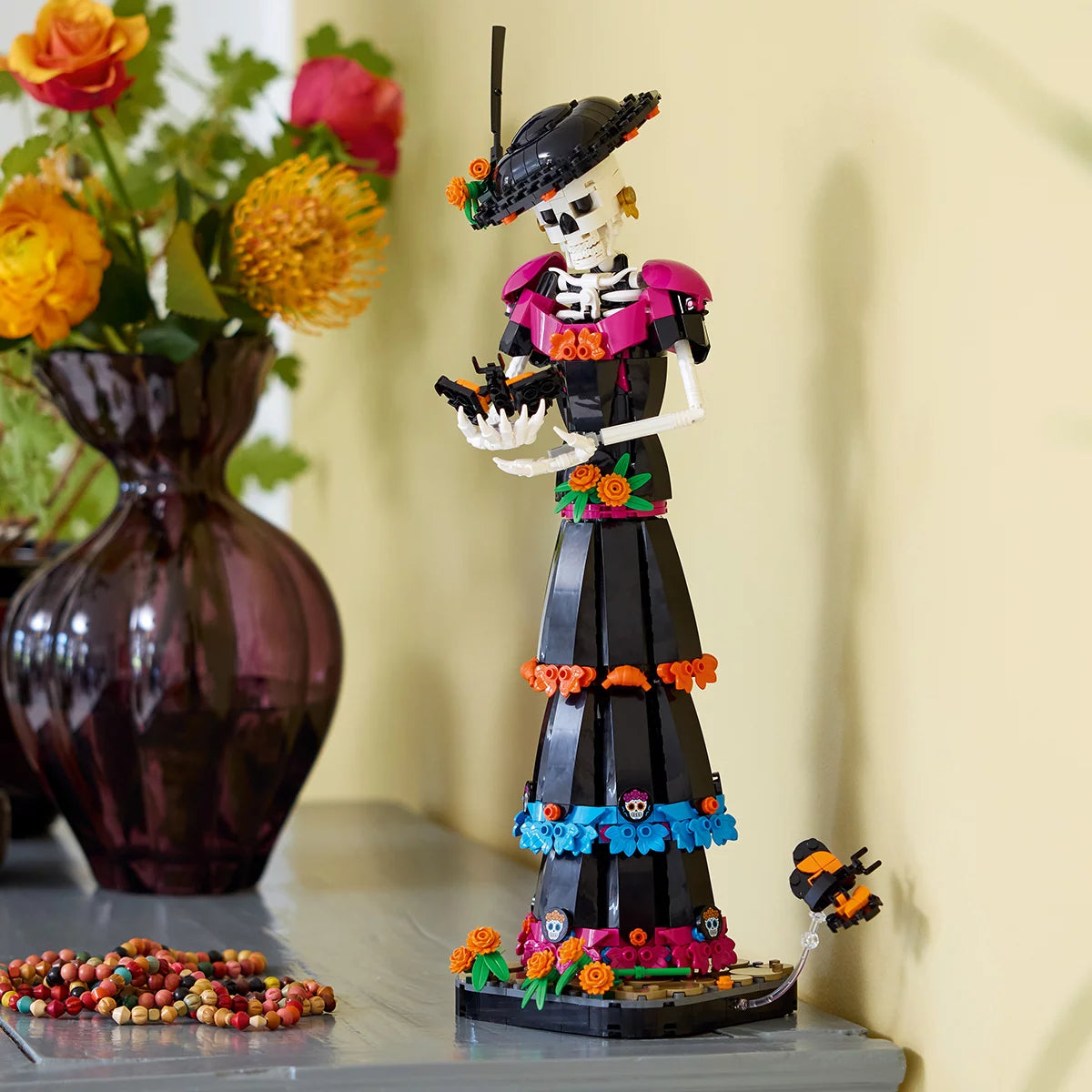 LEGO Ideas La Catrina model figure standing on a marigold-covered base wearing a wide-brimmed hat with colorful skull prints and orange flowers.