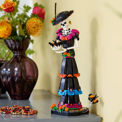LEGO Ideas La Catrina model figure standing on a marigold-covered base wearing a wide-brimmed hat with colorful skull prints and orange flowers.
