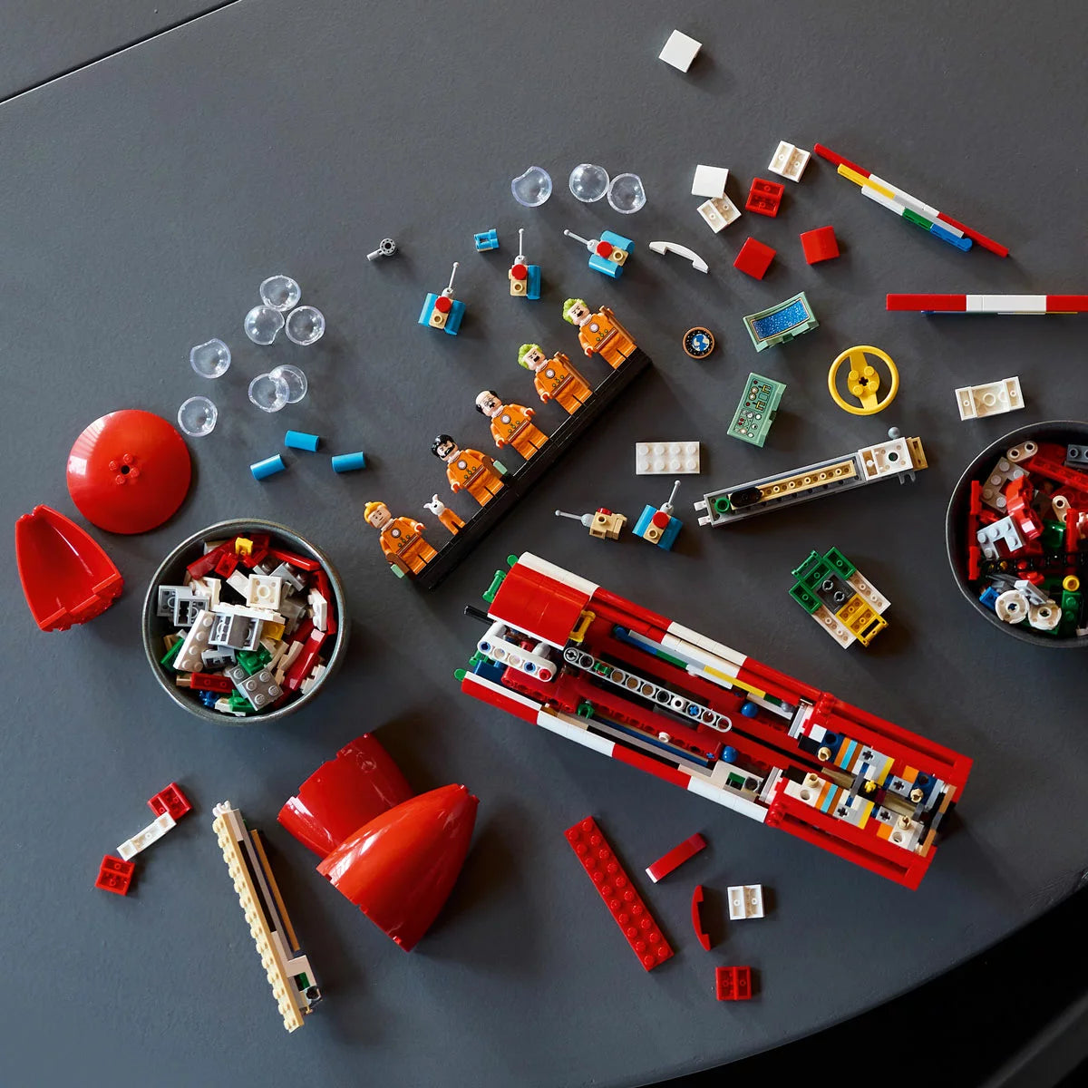 Profile view of the built tall red and white checkered LEGO 21367 Tintin Moon Rocket standing vertically next to five minifigures in orange spacesuits.