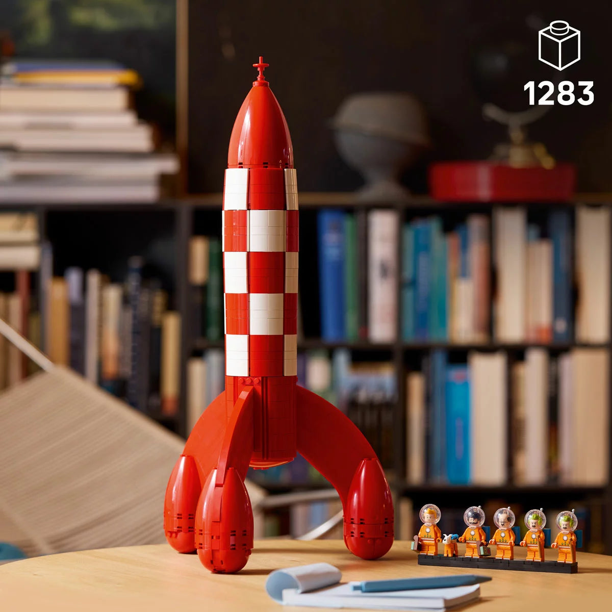 Profile view of the built tall red and white checkered LEGO 21367 Tintin Moon Rocket standing vertically next to five minifigures in orange spacesuits.