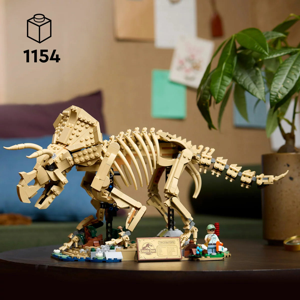 Side profile of the LEGO Jurassic World Triceratops Fossil 77985 skeletal model standing on a Cretaceous display base with ancient plants, an info plaque, and a Dr. Gerry Harding minifigure against a neutral dark backdrop.