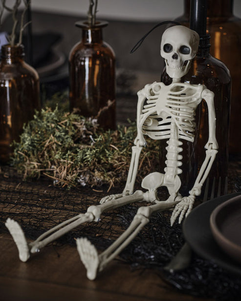 Full-length view of a 35cm off-white plastic hanging skeleton with posable joints suspended by a black rope against a rustic dark backdrop.
