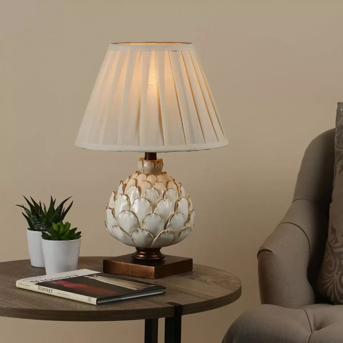 A close-up shot of the Layer table lamp showing its textured cream artichoke resin base on a mottled gold wood plinth, topped with a pleated ecru shade in a cozy country cottage bedroom.
