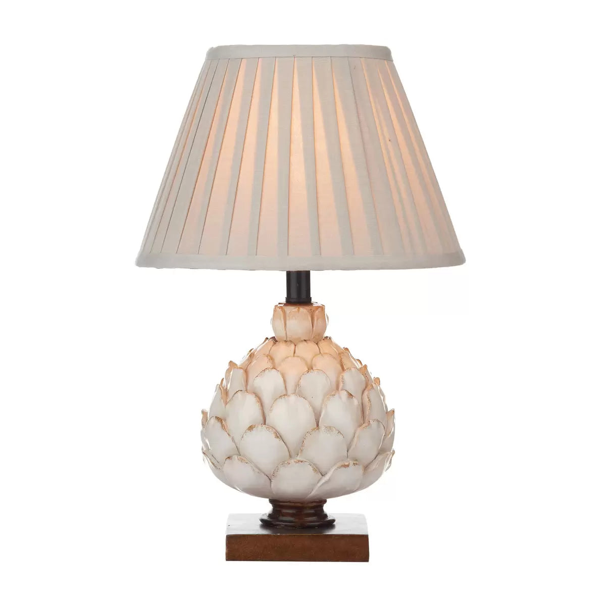 A close-up shot of the Layer table lamp showing its textured cream artichoke resin base on a mottled gold wood plinth, topped with a pleated ecru shade in a cozy country cottage bedroom.