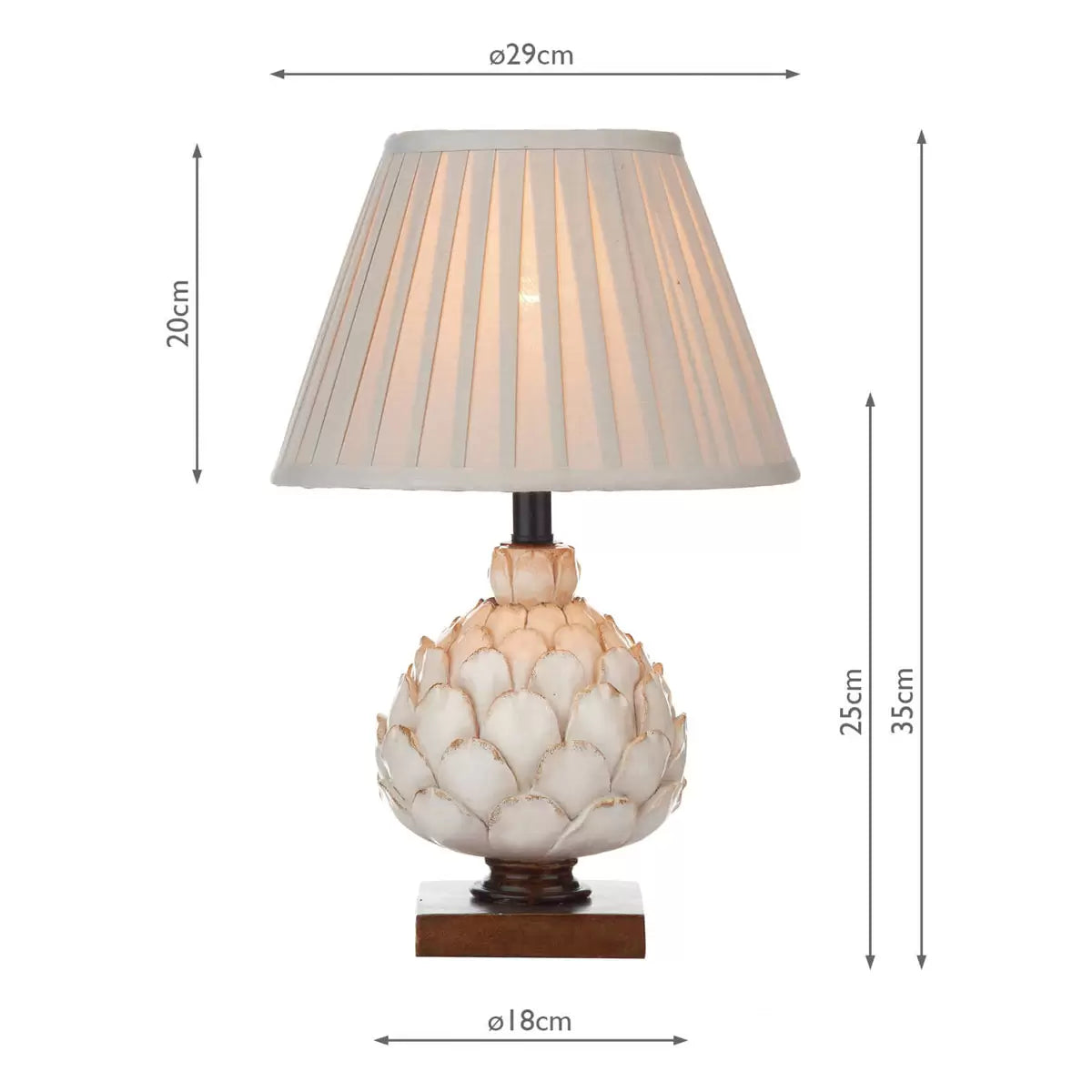 A close-up shot of the Layer table lamp showing its textured cream artichoke resin base on a mottled gold wood plinth, topped with a pleated ecru shade in a cozy country cottage bedroom.