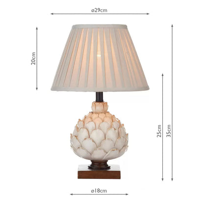 A close-up shot of the Layer table lamp showing its textured cream artichoke resin base on a mottled gold wood plinth, topped with a pleated ecru shade in a cozy country cottage bedroom.