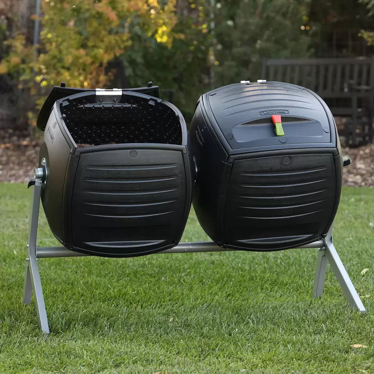 A side-by-side view of the Lifetime 378L dual tumbling composter on a sturdy steel base, situated on a clean garden patio, showing the black panels and red processing indicators.