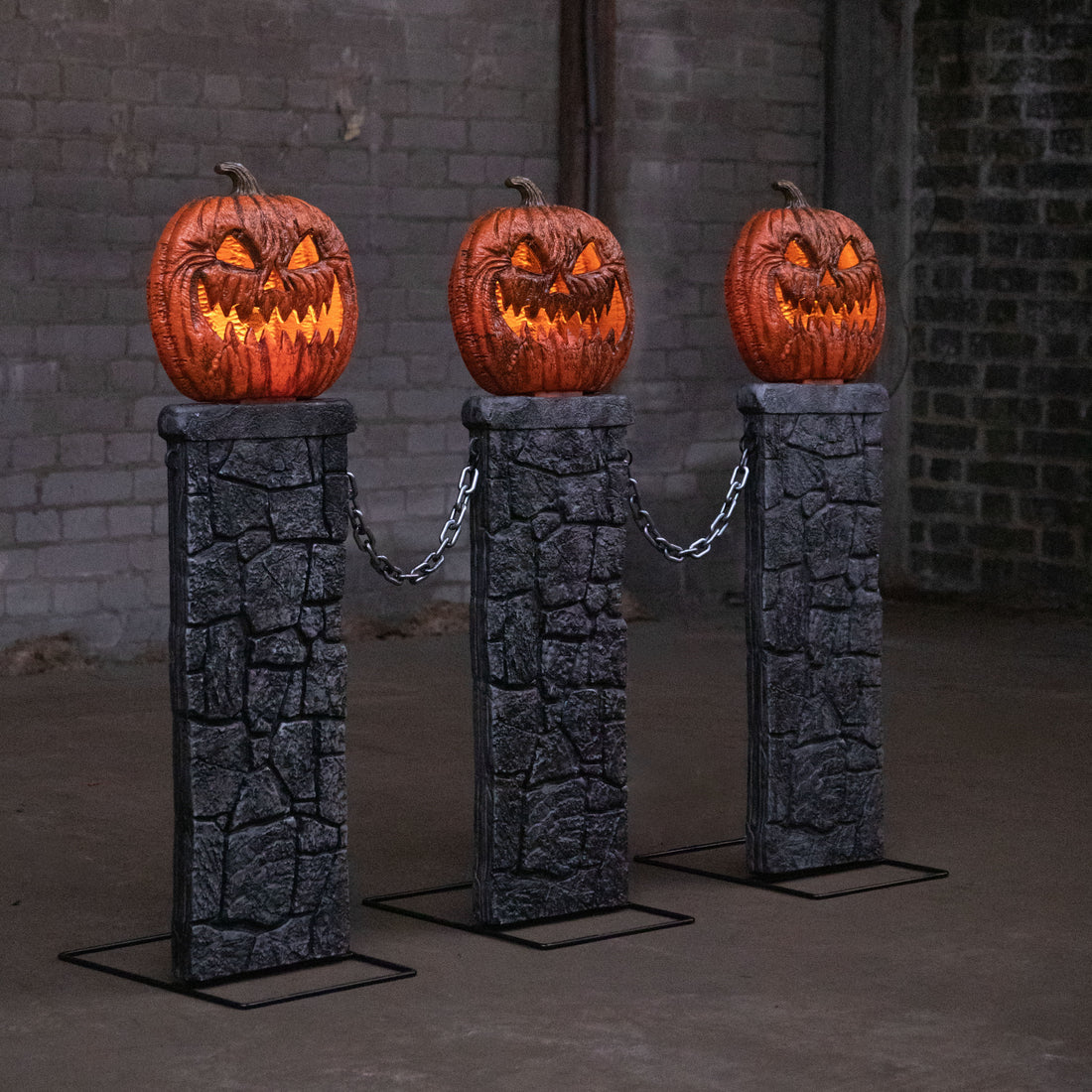 Three grey gothic stone pedestals with orange lit LED jack-o'-lantern tops connected by black chains on a dark outdoor lawn.