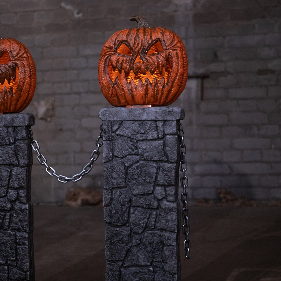 Three grey gothic stone pedestals with orange lit LED jack-o'-lantern tops connected by black chains on a dark outdoor lawn.