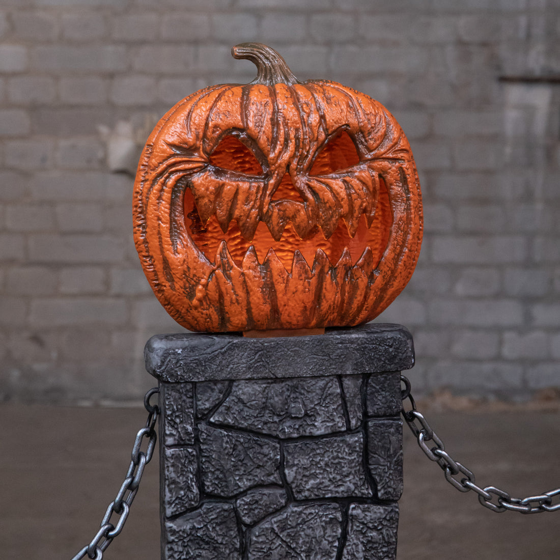 Three grey gothic stone pedestals with orange lit LED jack-o'-lantern tops connected by black chains on a dark outdoor lawn.