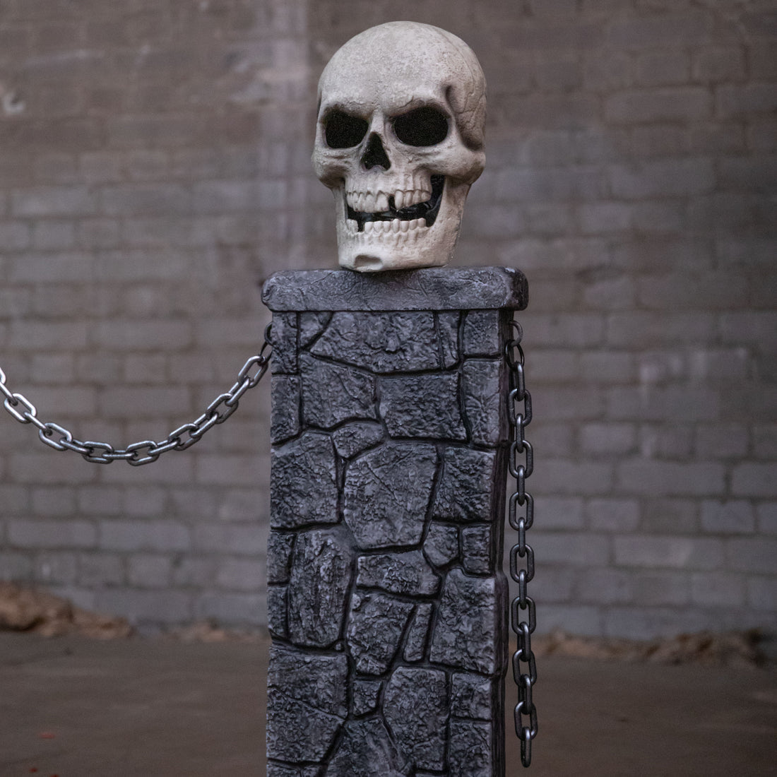 Samuel Alexander grey stone-effect Halloween skull fence with three illuminated LED pedestals and connecting chains set up on a dark lawn.