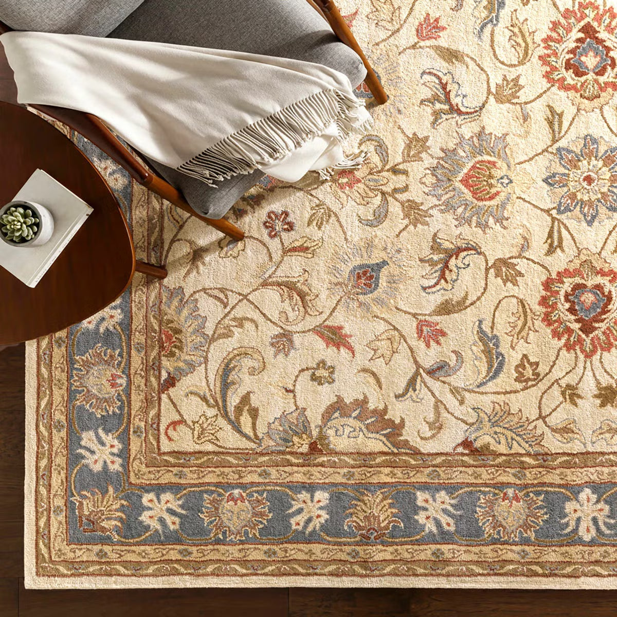 Top-down angle showing the detailed cream and muted floral pattern of the hand-tufted Livabliss Caesar wool rug styled on light oak flooring under a wooden coffee table.