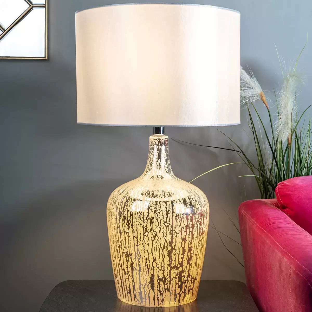 A Lolek silver glass table lamp with a glowing, textured mercury finish base and an ivory faux silk drum shade illuminating a cozy bedside table environment.