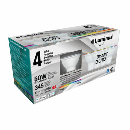 Four white Luminus Smart GU10 LED spotlight bulbs arranged neatly on a dark wooden countertop casting a soft multi-coloured glow.