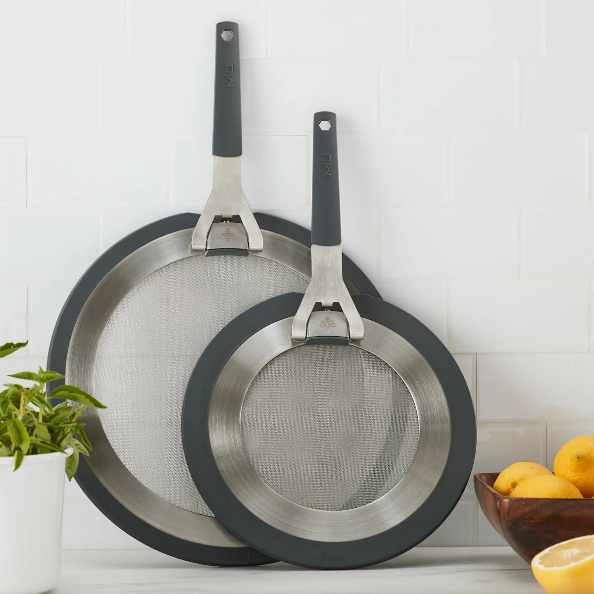 MIU 2-piece stainless steel splatter screen set with foldable silicone handles displayed next to frying pans.