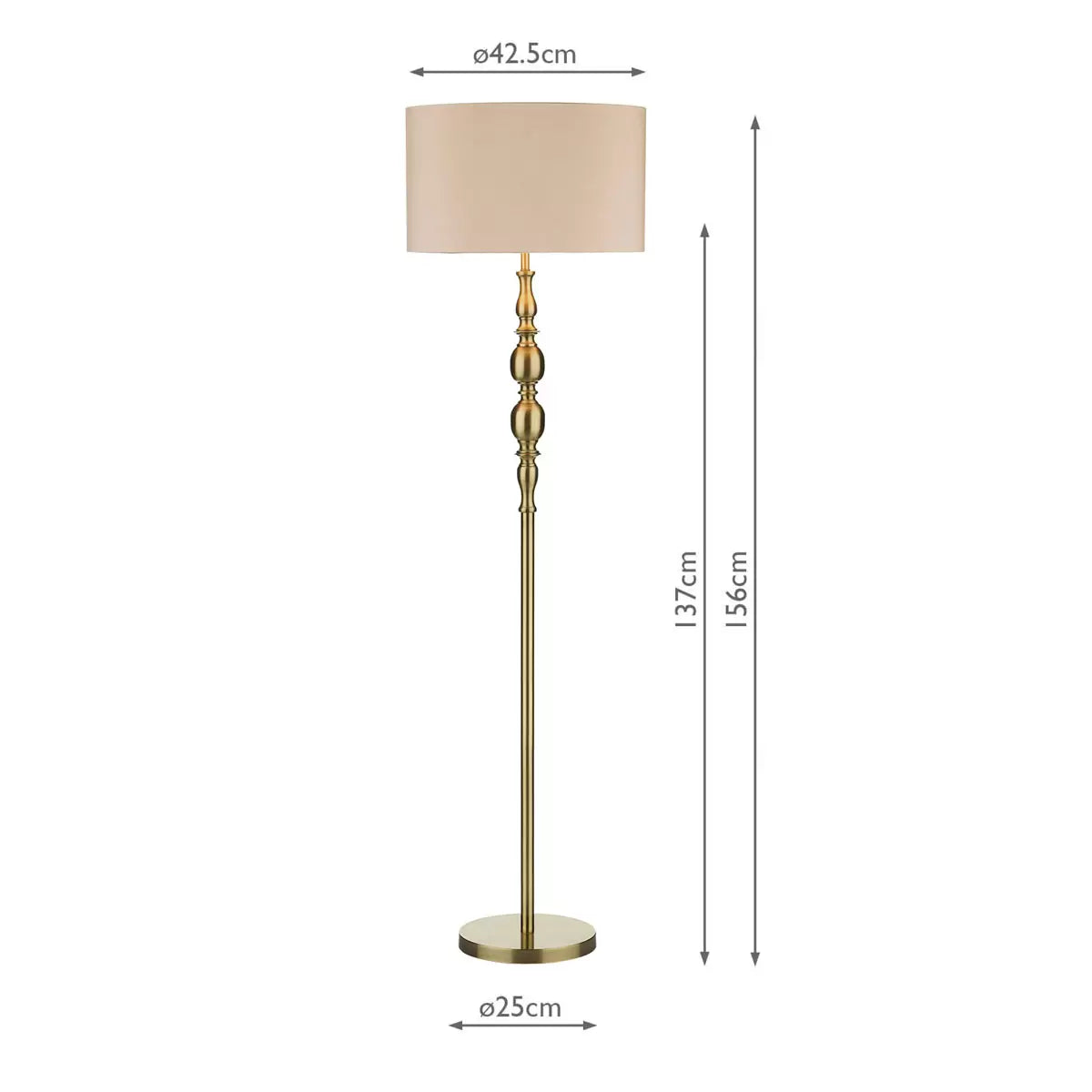 A tall Madrid antique brass metal floor lamp with a beige faux silk drum shade stands beside a grey armchair in a warmly lit living room setting.