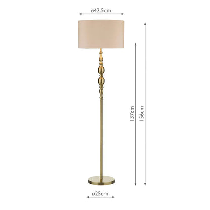 A tall Madrid antique brass metal floor lamp with a beige faux silk drum shade stands beside a grey armchair in a warmly lit living room setting.