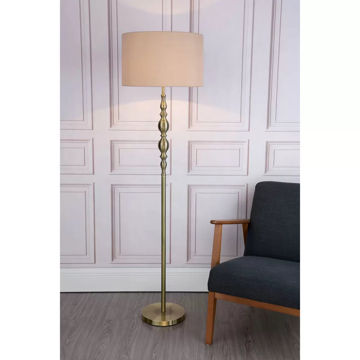 A tall Madrid antique brass metal floor lamp with a beige faux silk drum shade stands beside a grey armchair in a warmly lit living room setting.