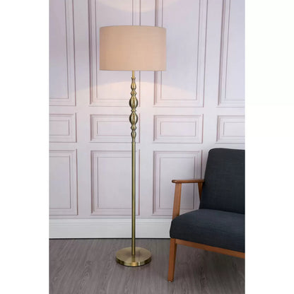 A tall Madrid antique brass metal floor lamp with a beige faux silk drum shade stands beside a grey armchair in a warmly lit living room setting.