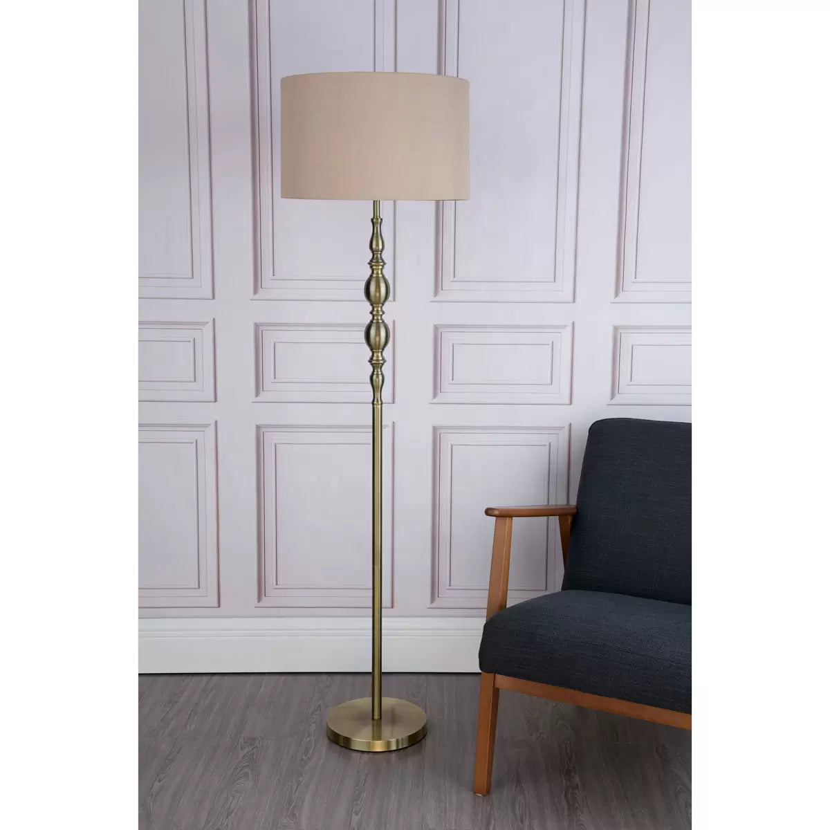 A tall Madrid antique brass metal floor lamp with a beige faux silk drum shade stands beside a grey armchair in a warmly lit living room setting.