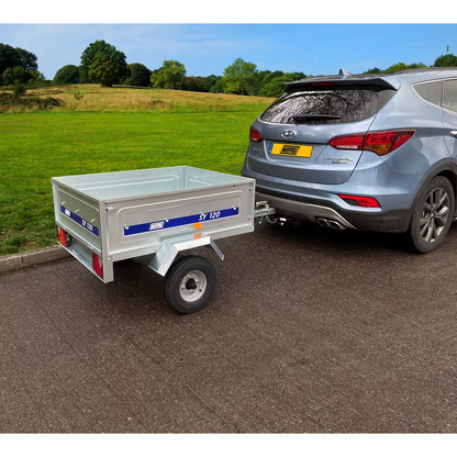 A silver galvanised steel Maypole SY120 utility trailer with its grey weather-resistant PVC flat cover tied down tightly with elasticated cords, viewed from a three-quarter angle.