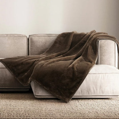 A plush grey Mon Chateau faux fur throw draped casually over the arm of a dark leather sofa in a cozy living room setup.